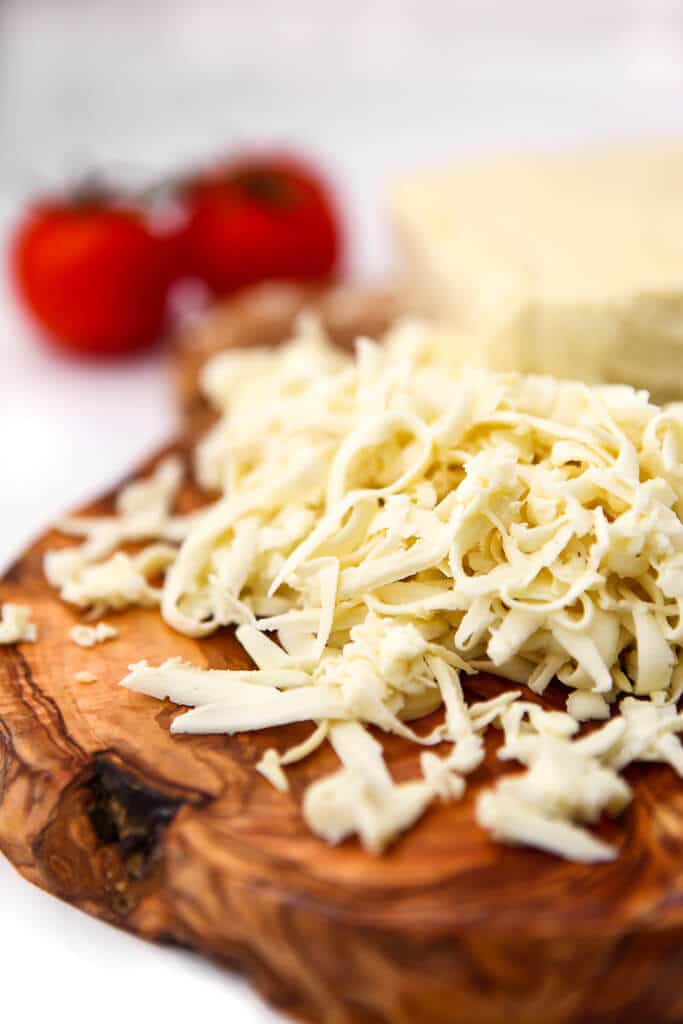 Shredded vegan mozzarella cheese on a wooden cutting board with two tomatoes behind it. 