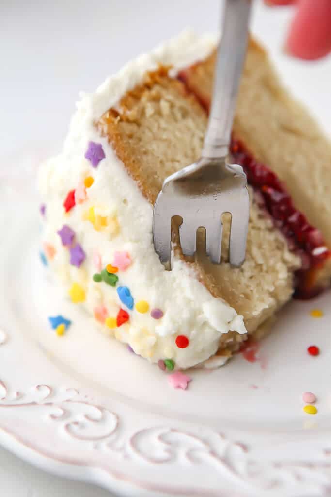 A slice of vegan vanilla cake with vanilla frosting, sprinkles, and a jam layer in the middle.