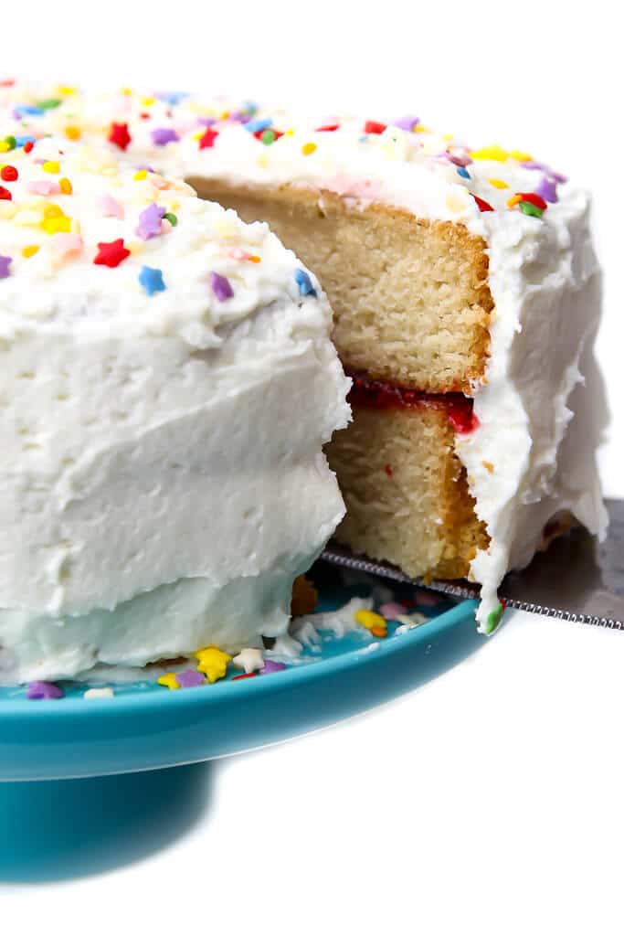 A vegan white cake with vanilla frosting with a slice being taken out of it.