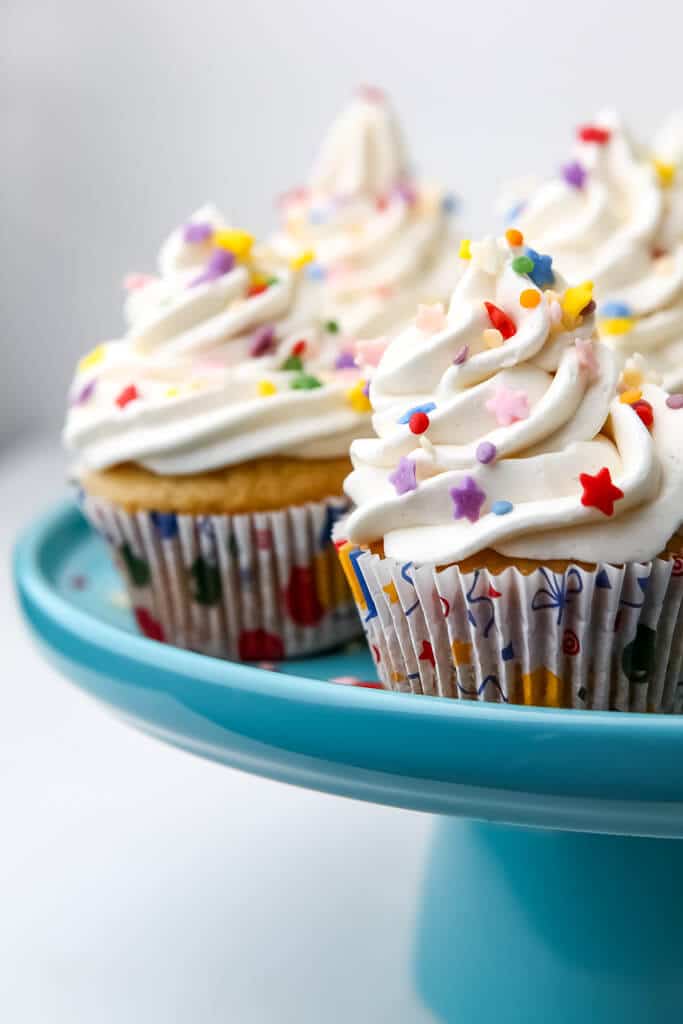 Vegan vanilla cupcakes with vanilla frosting and colorful sprinkles on a blue cake dish.