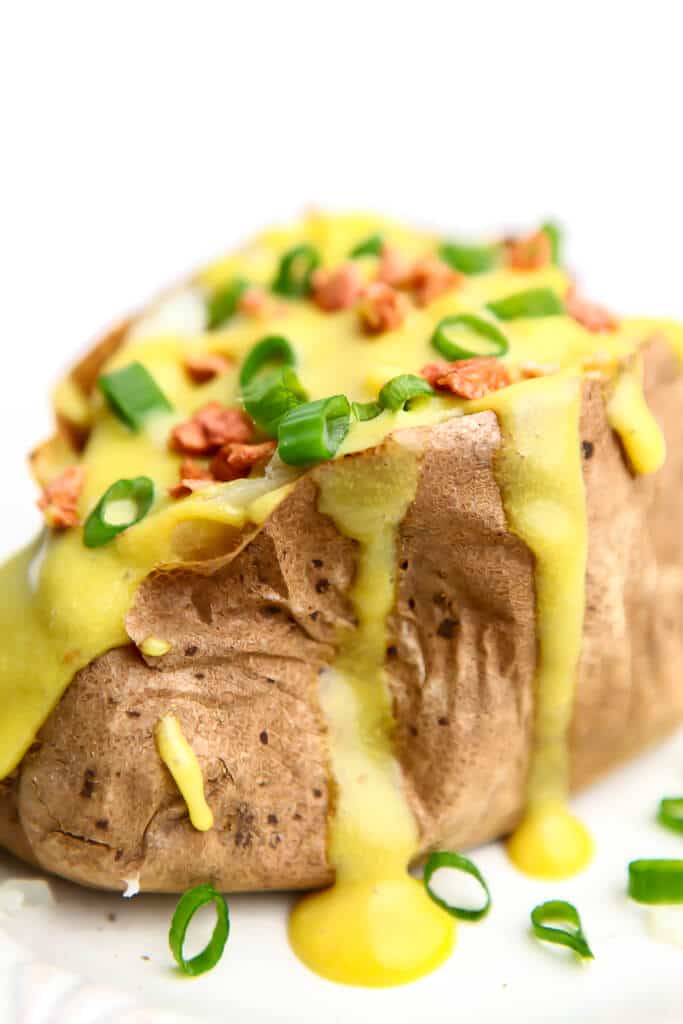 A baked potato with vegan nacho cheese sauce, vegan bacon bits, and green onions on top.