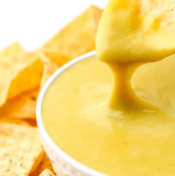 A corn chip being dipped into a bowl of vegan nacho cheese sauce with cheese dripping off of it.