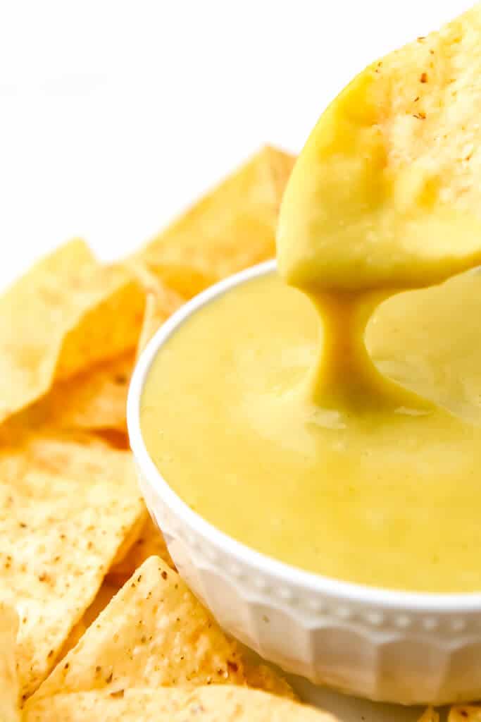 A corn chip being dipped into a bowl of vegan nacho cheese sauce with cheese dripping off of it.