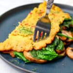 A chickpea omelette filled with vegan cheese, spinach, and mushrooms on a blue plate with a fork sticking in it.