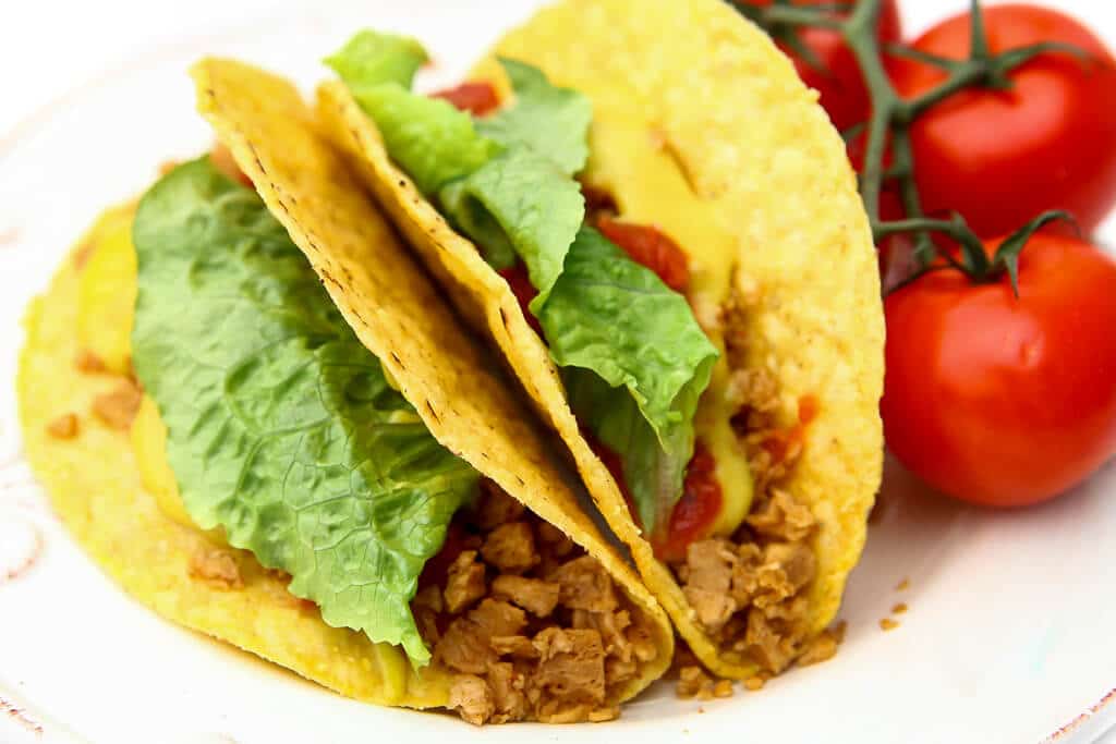 Two vegan tacos with TVP taco meat, lettuce, vegan nacho cheese, and tomatoes on a white plate.