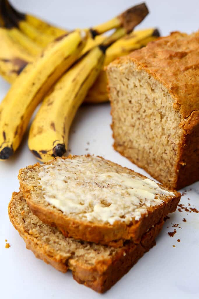 A loaf of vegan banana bread with two slices taken out of it and bananas behind it.