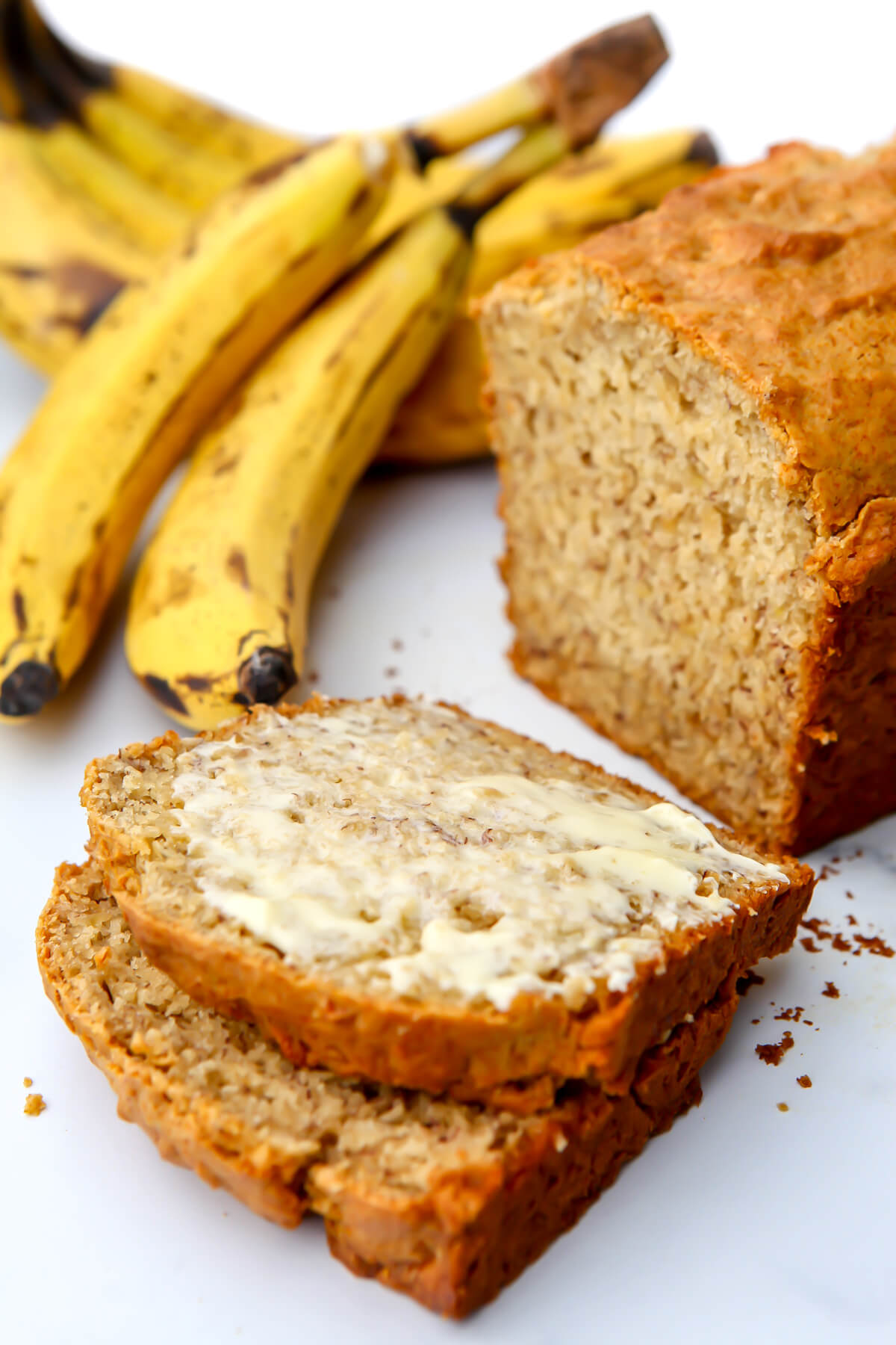 A loaf of vegan banana bread with two slices taken out of it and bananas behind it.