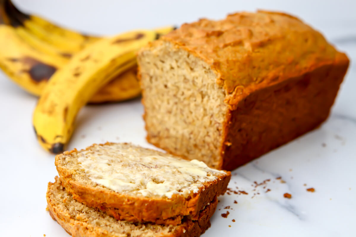 Vegan banana bread on a marble counter top sliced with some bananas behind it.