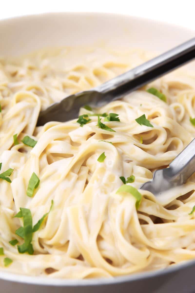 A pan full of coconut Alfredo sauce being mixed with fettuccine noodles using tongs.