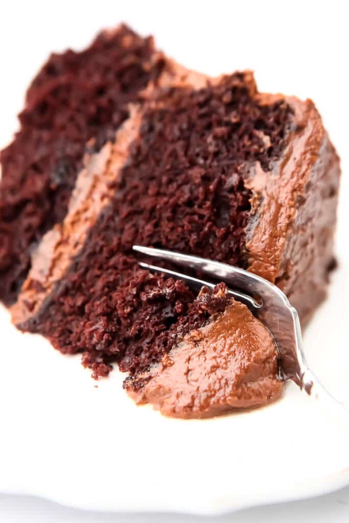A slice of vegan chocolate cake with chocolate frosting on a white plate with a fork cutting a piece off of it.
