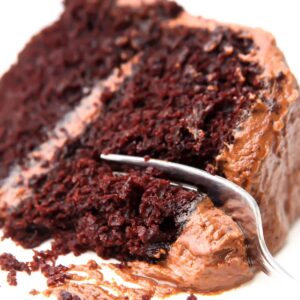 A slice of vegan chocolate cake with chocolate frosting on a white plate with a fork cutting a piece off of it.