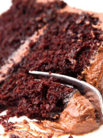 A slice of vegan chocolate cake with chocolate frosting on a white plate with a fork cutting a piece off of it.