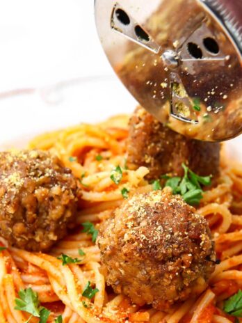 A plate of vegan spaghetti and meatballs with a silver shaker sprinkling vegan Parmesan cheese over the top.