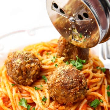 A plate of vegan spaghetti and meatballs with a silver shaker sprinkling vegan Parmesan cheese over the top.