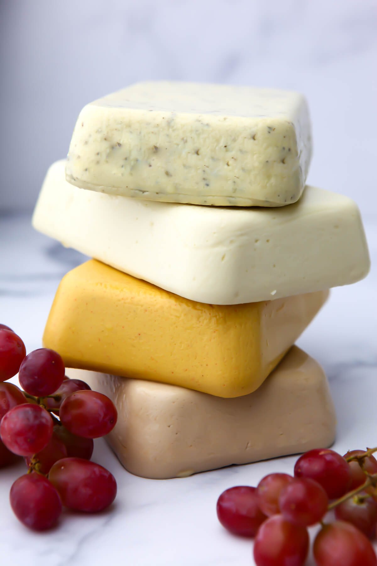 A stack of 4 flavors of vegan cheese with red grapes next to them.