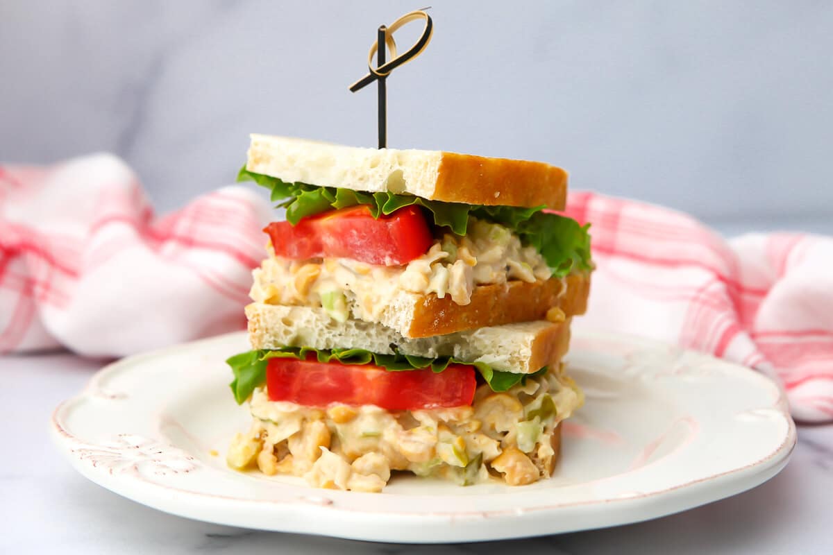 A chickpea salad sandwich on a white plate with a red and white towel behind it.