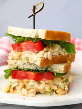 A vegan chickpea salad sandwich with lettuce and tomato slices on it on a white plate with a red tea towel behind it.