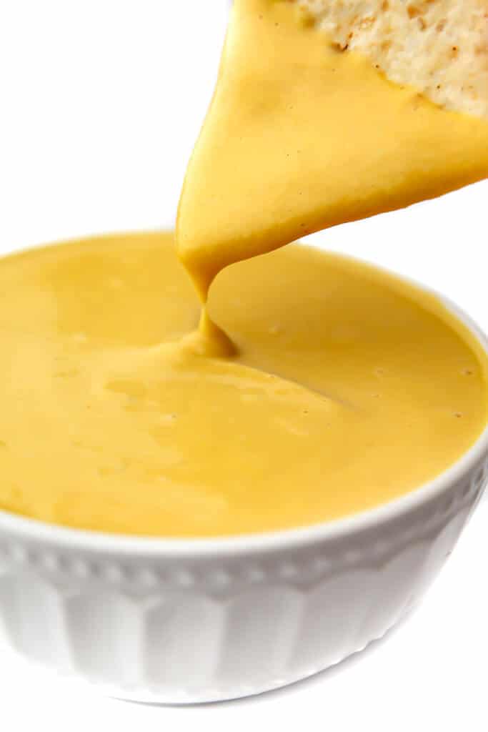 A vegan cheese sauce made from vegetables in a white bowl with a chip being dipped into it.