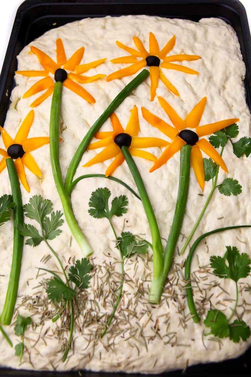 Focaccia bread decorated with yellow peppers, black olives, and herbs to form sunflowers.