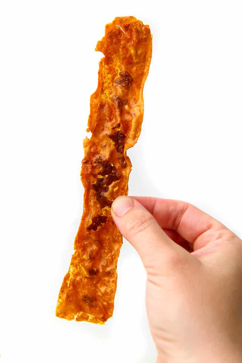 A slice of crispy vegan bacon being held in the air with a white background.