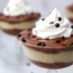 A close up of a glass dish layered with vegan chocolate and vanilla pudding with whipped cream and mini chocolate chips on top.