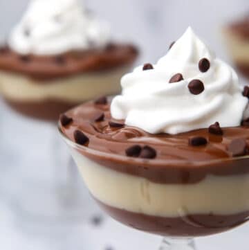 A close up of a glass dish layered with vegan chocolate and vanilla pudding with whipped cream and mini chocolate chips on top.