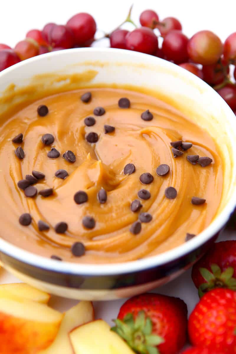 A bowl of peanut butter dip with mini chocolate chips sprinkled on top and fruit around it.
