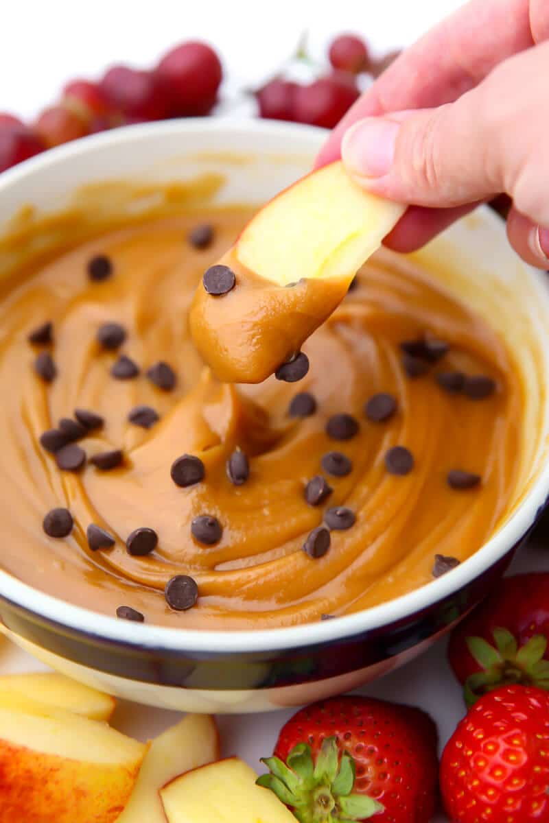 A bowl of peanut butter dip with chocolate chips on it with an apple slice being dipped into it.