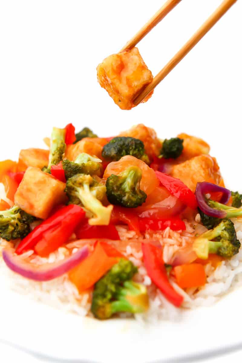 A plate of vegan sweet and sour tofu with mixed veggies with a piece of tofu held in chop sticks.