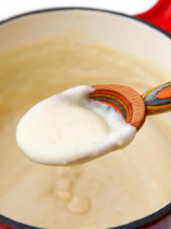 A colorful wooden spoon filled with vegan Bechamel sauce over a saucepan.