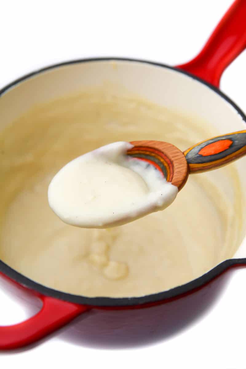A red saucepan with vegan Bechamel sauce on a colorful wooden spoon.