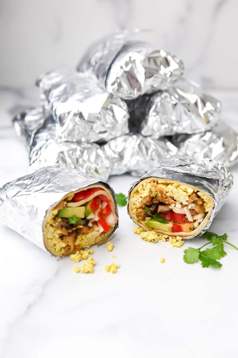 A stack of vegan breakfast burritos wrapped in foil to be frozen with 1 burrito in front cut in half so you can see the contents.