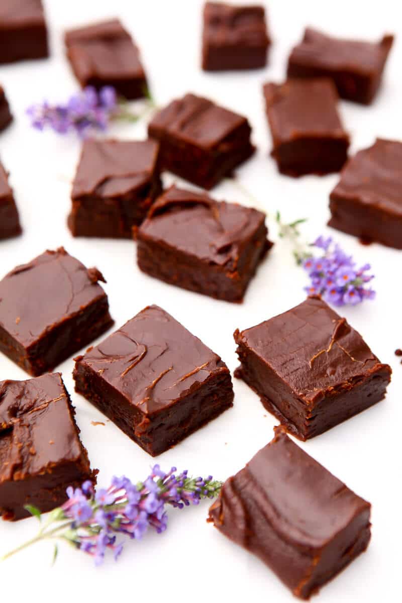 Many squares of vegan fudge scattered around on top of white parchment paper with some purple flowers around.