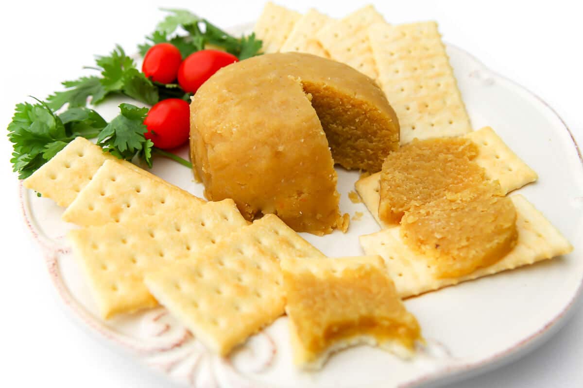 A round of cheese made from almonds on a white plate with crackers, greens, and cherry tomatoes.