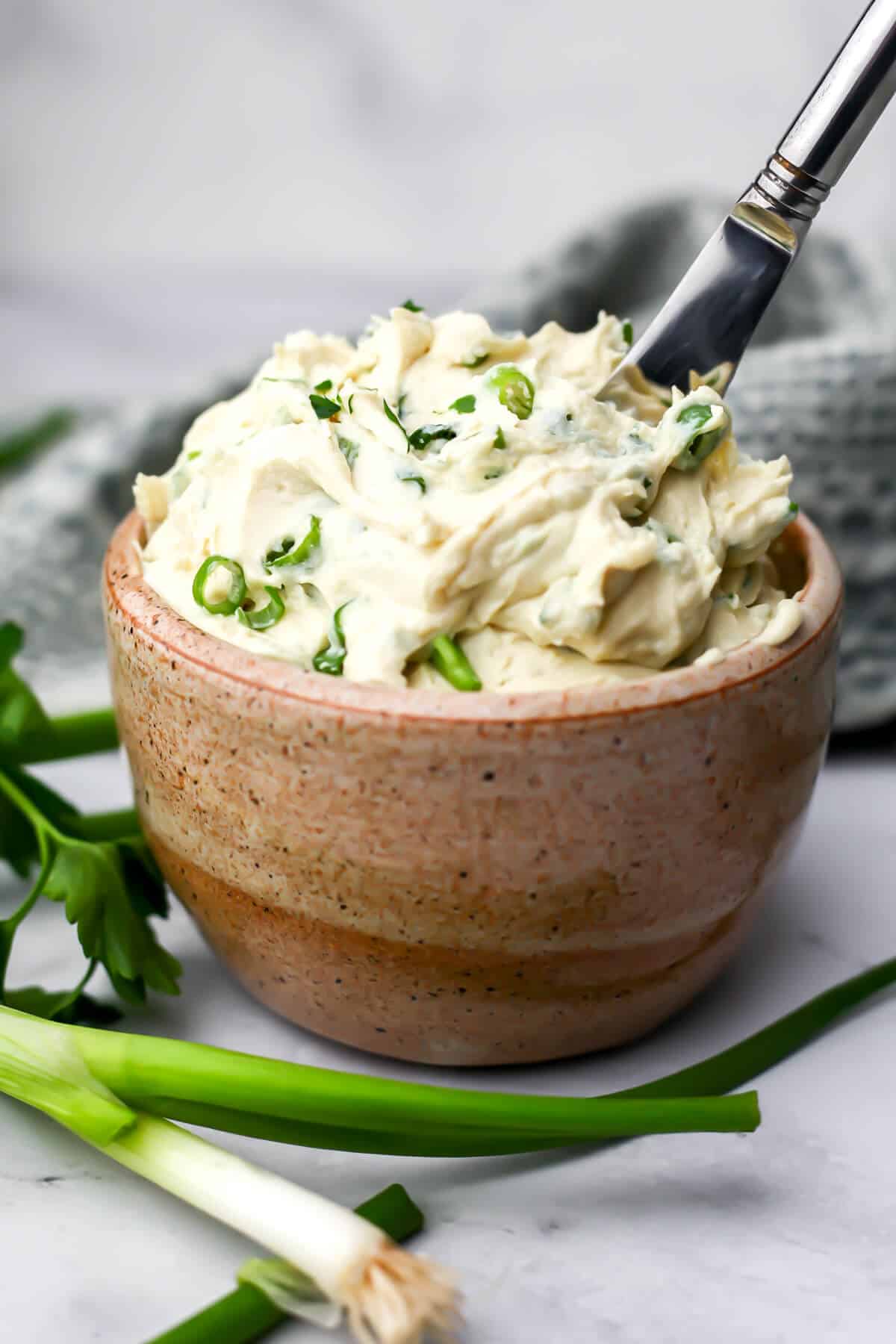 A bowl of vegan garlic herb cream cheese with a knife in it and herbs around it.