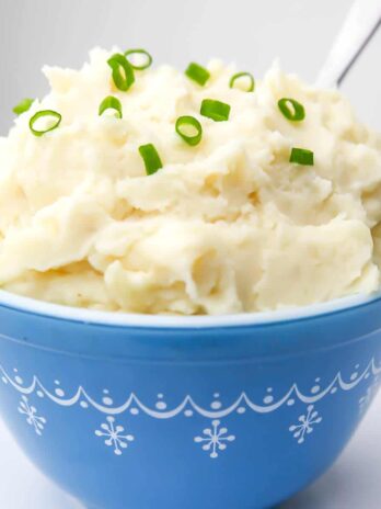 A blue bowl filled with vegan mashed potatoes with chives on top and a spoon stuck in the side.