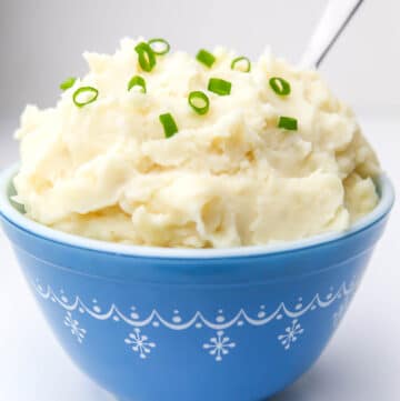 A blue bowl filled with vegan mashed potatoes with chives on top and a spoon stuck in the side.