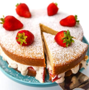 A vegan sponge cake with powdered sugar and strawberries on top with a slice being taken out of it.