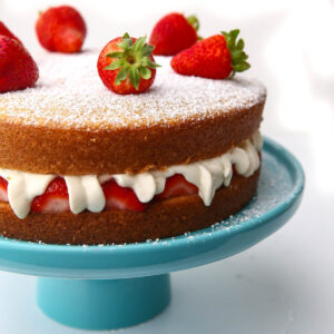 A vegan Victorian sponge cake with whipped cream and strawberries in the middle on a blue cake dish.