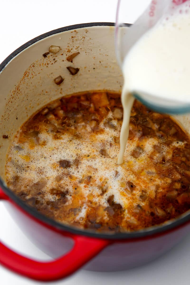 Vegan milk being pour into a red sauce pan filled with sauted mushrooms.