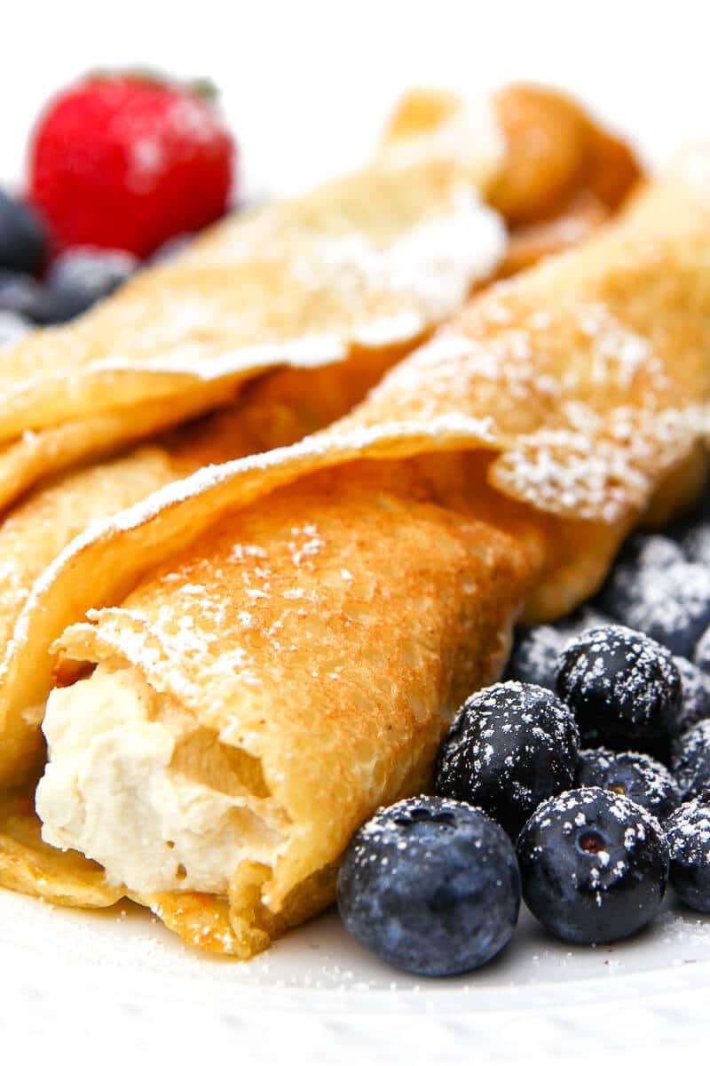 Vegan crepes with tofu ricotta filling and blueberries and strawberries on the side dusted with powdered sugar.