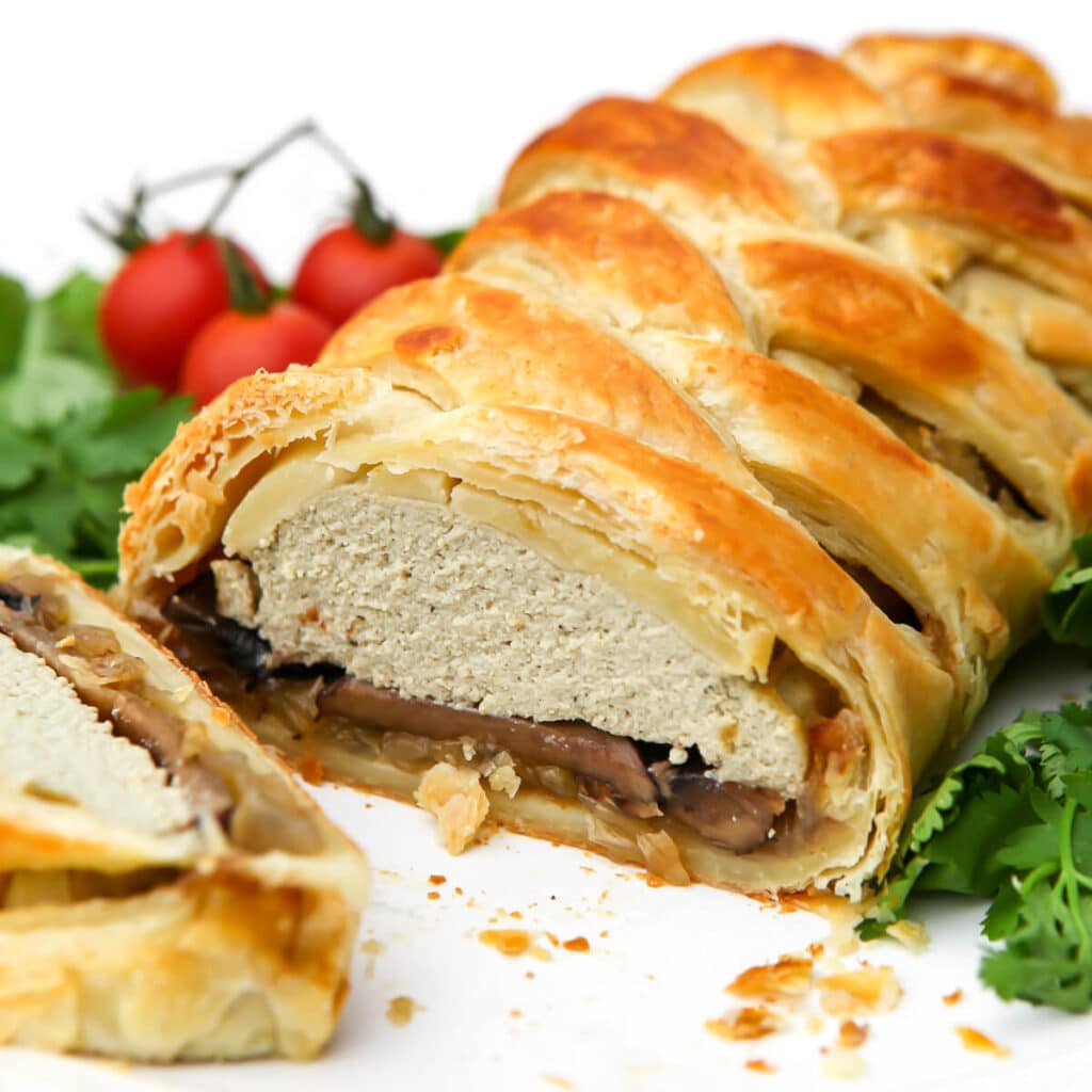 A vegan wellington filled with onions, mushrooms, and tofu on a white plate with a slice cut out of it.