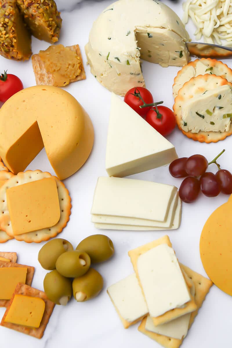 A variety of vegan cheeses like smoked gouda, cheddar, and tofu herb cheese on a marble cutting board.
