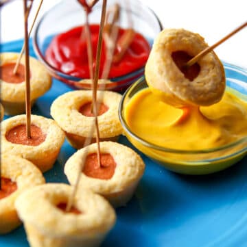 A blue plate filled with mini vegan corn dog bites with one being dipped into a bowl of mustard.