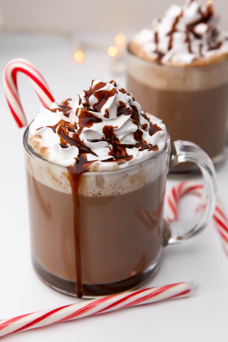 Two cups of vegan hot cocoa with whipped cream and vegan chocolate syrup on top and candy canes around.. 