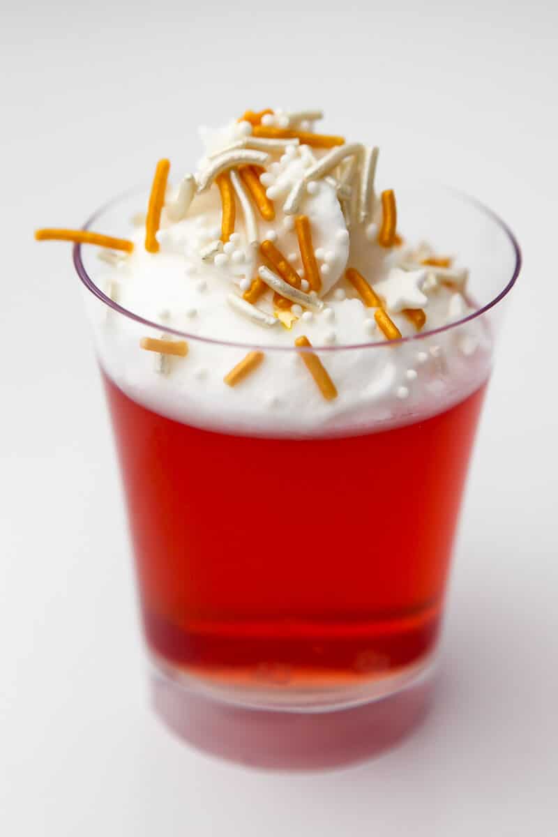 A single shot glass filled with vegan jello mixed with vodka topped with whipped cream and sprinkles.