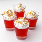 Four little plastic shot glasses filled with vegan jello shots topped with vegan whipped cream and sprinkles.