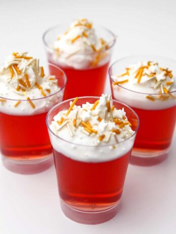 Four little plastic shot glasses filled with vegan jello shots topped with vegan whipped cream and sprinkles.