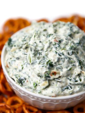 A close up of vegan spinach dip in a white bowl with pretzels around it.
