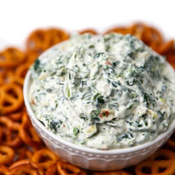 A close up of vegan spinach dip in a white bowl with pretzels around it.
